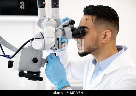 Young Arab Dentist Doctor Using Dental Microscope At Workplace In Clinic, Side View Shot Of Middle Eastern Stomatologist Making Research With Modern M Stock Photo