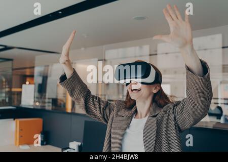 Young excited redhead businesswoman in formal wear sitting in office interior using VR headset