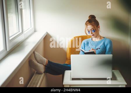 Front view of an young woman studying from home at the computer, sitting on armchair making notes and listening to music Stock Photo