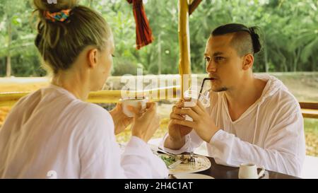 Man and woman eat and drink tasty beverages spending time in traditional local floating cafe on water. Loving happy couple having breakfast outdoors Stock Photo