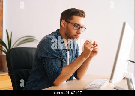 Side view of serious handsome young freelance designer male in stylish glasses working on desktop computer holding pen in hands Stock Photo