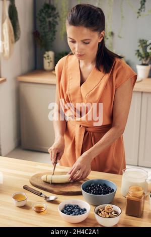 Young woman chopping peeled banana on wooden board while preparing ingredients for homemade smoothie or yoghurt Stock Photo