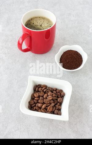 Whole and ground coffee beans in white bowls and black coffee in a red mug on gray concrete. High angle view. Stock Photo