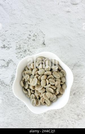 Green whole coffee beans in white bowl on rough textured white concrete background with copy space. Top view. Vertical version. Stock Photo