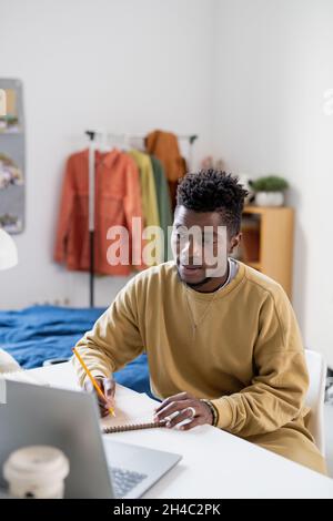 African guy in casualwear looking at laptop display while making notes during online lesson at home Stock Photo