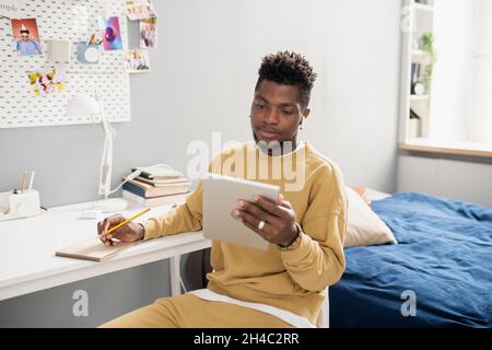 Young serious student in casualwear looking at tablet screen and making notes in notepad during online lesson Stock Photo