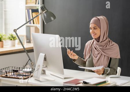 Happy young Muslim woman talking to teacher on computer screen during online lesson in office environment Stock Photo
