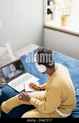 Young student in headphones and homewear listening to teacher and making notes while sitting on bed during online lesson Stock Photo