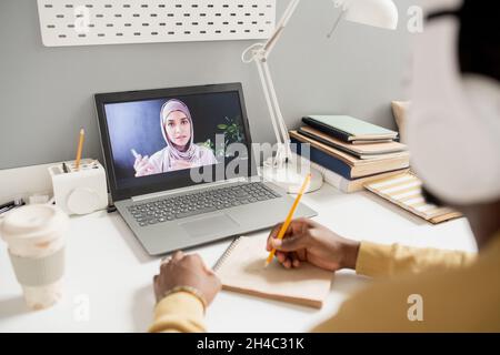 Young Muslim woman on laptop screen teaching online learners while contemporary African student making notes Stock Photo