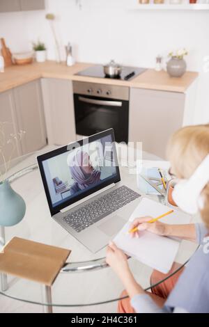 Mature student making notes while listening to teacher with paper on laptop screen during online course of study Stock Photo