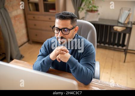 Serious middle-aged teacher or student in eyeglasses sitting in front of computer screen and taking part in online lesson Stock Photo