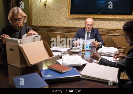 Three contemporary lawyers working with papers while two of them reading contracts and mature woman looking for documents in box Stock Photo