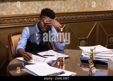 Young African male lawyer in formalwear touching his head while looking through juridical documents by workplace Stock Photo