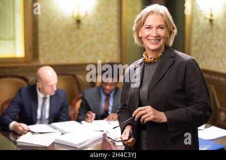 Happy experienced female lawyer in formalwear standing in front of camera with two colleagues working with documents on background Stock Photo