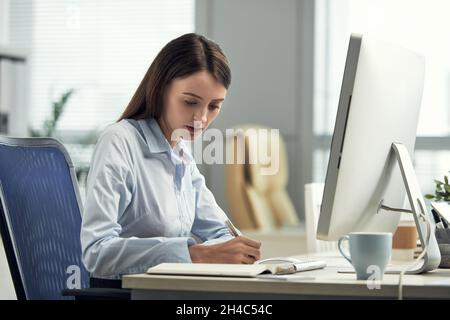 Concentrated young Caucasian businesswoman making notes in diary while planning work day in modern office Stock Photo