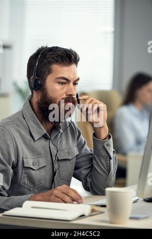 Serious young bearded call center operator in headset with microphone sitting at desk and using laptop Stock Photo