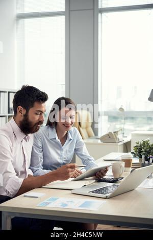 Team of young Caucasian managers sitting at office table and using tablet while working on new project Stock Photo