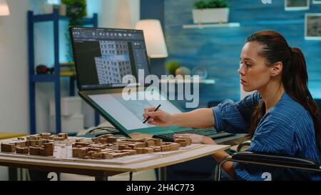 Woman with disability using touch screen for construction layout at office. Architect in wheelchair analyzing building model on computer and table, creating blueprint plan for development Stock Photo