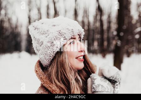 Beautiful young woman in snowy fancy winter woodland. Girl wearing fluffy gloves, cap and coat. Christmas forest, trees on blurred background. Crossed Stock Photo