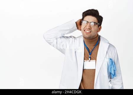 Clueless doctor scratching head and looking confused up, cant understand. Puzzled intern standing in medical uniform, looking aside, white background Stock Photo