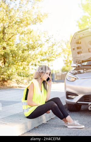Woman sitting on the curb calling her insurance company about a breakdown in her car Stock Photo