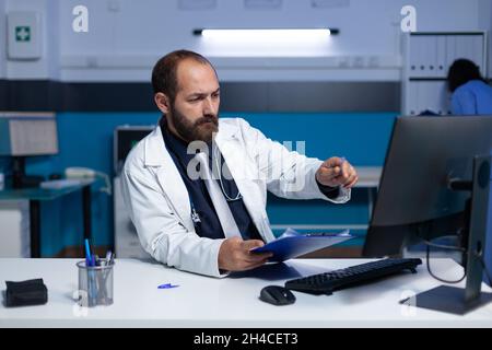 Doctor using computer and checkup files for healthcare work at night. Specialist analyzing information on monitor for prescription medicine and medical advice, working late at office. Stock Photo