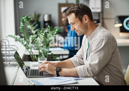 Side view portrait of smiling adult man using laptop while enjoying work in office, copy space Stock Photo