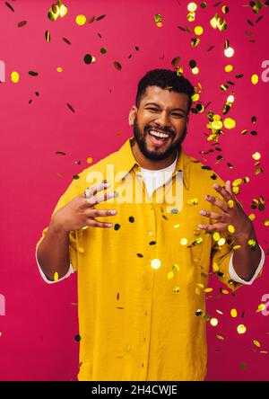 Golden confetti in the studio. Excited young man smiling cheerfully while standing under falling confetti in a studio. Happy young man celebrating lif Stock Photo