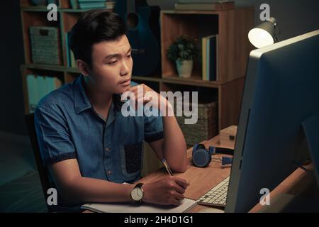 Serious young Asian man sitting at wooden table and making notes in sketchpad while working with computer late at home Stock Photo