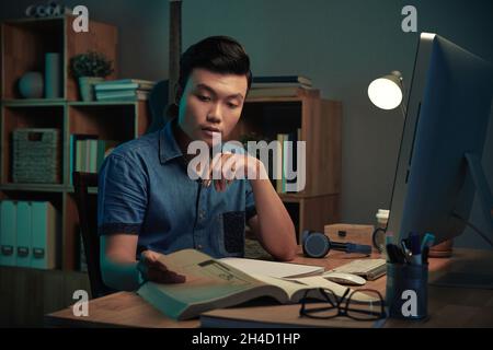 Serious young Asian programmer sitting under lamp light at desk and reading manual at night Stock Photo