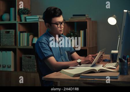Serious young Vietnamese programmer in eyeglasses using digital tablet in dark home office Stock Photo