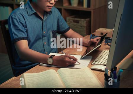 Close-up of serious busy Asian app developer sitting at table with computer and manual and writing out code in sketchpad Stock Photo