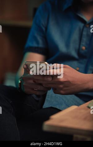Close-up of unrecognizable man in casual shirt sitting at table and texting message on smartphone in dark room Stock Photo