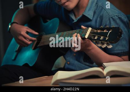 Close-up of young man in casual shirt sitting at table with open book and playing guitar in darkness Stock Photo