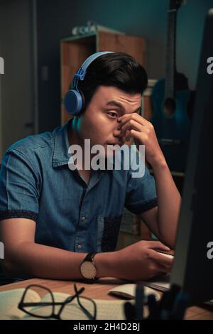 Tired young Asian programmer in wireless headphones sitting at desk and rubbing bridge of nose while working late Stock Photo