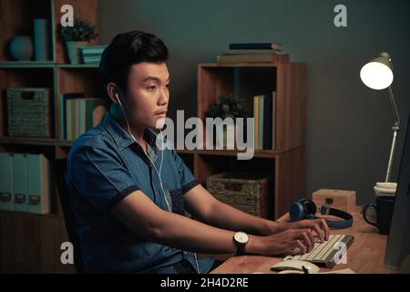 Serious focused young Asian designer in earphones sitting at desk and typing on computer keyboard in dark room Stock Photo