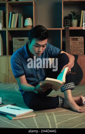 Concentrated young Asian man sitting on carpet at home and reading textbook in dark room Stock Photo