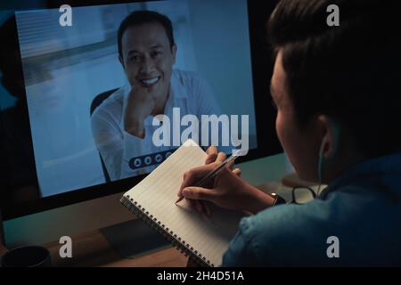 Over shoulder view of Asian man making notes in sketchpad while listening to boss during online meeting Stock Photo