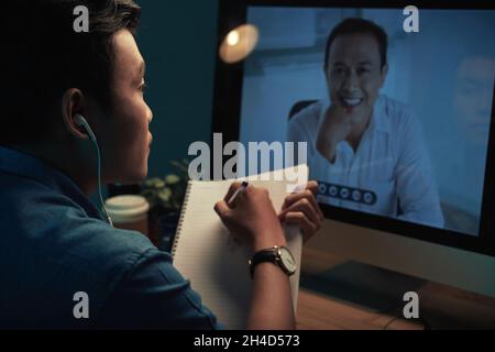 Over shoulder view of Asian student in earphones watching webinar and making notes in sketchpad at night Stock Photo