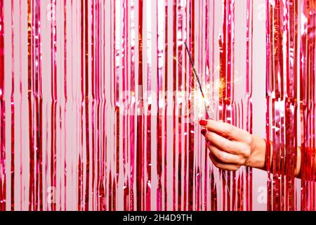 Woman hand with nail polish in red breaking through a pink tinsel with a burning sarkler. Party concept Stock Photo