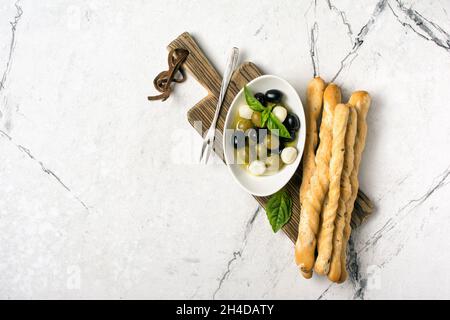 Top view of grissini with basil spice, green and black olives in oil, mozzarella on wooden board and white marble background with copy space Stock Photo