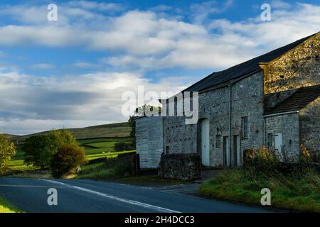 Old stone barn at side of quiet countryside B6160 road, rolling hills & sheep in sunlit farm fields under blue sky - Yorkshire Dales, England, UK. Stock Photo