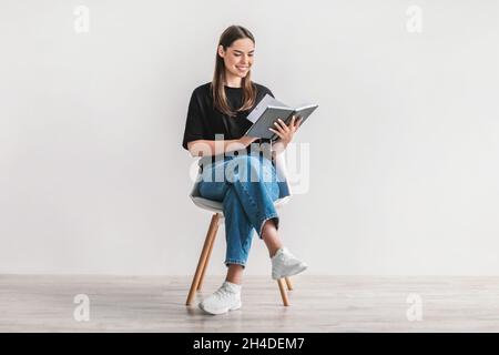 Stay home hobbies. Positive young woman reading favorite book, sitting on chair against white studio wall, full length Stock Photo