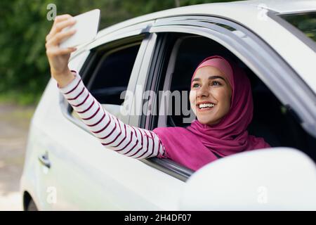 Joyful Muslim Woman In Hijab Taking Selfie While Sitting In Car Stock Photo