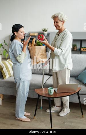 Vertical full length portrait of female caregiver bringing groceries to senior woman Stock Photo