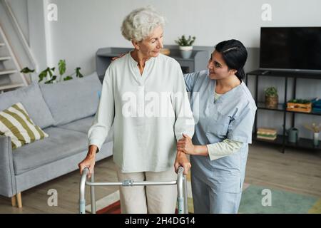 Waist up portrait of smiling female nurse helping senior woman using mobility walker in retirement home, copy space Stock Photo