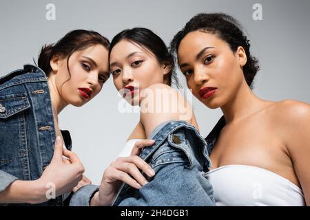 Low angle view of interracial women with red lips looking at camera isolated on grey