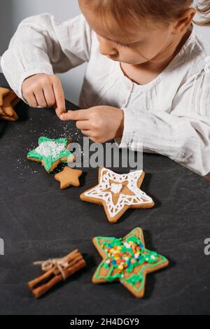 Child cooking and eating home made gingerbread cookies, stars, man. Happy toddler girl celebrated Christmas eve at home, Kid decorating pastry with Stock Photo