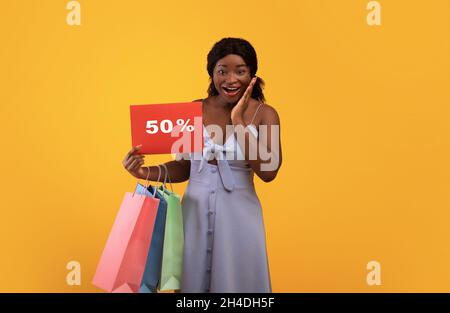 Emotional young black woman holding bright shopping bags and FIFTY PERCENT OFF sign, looking at camera in excitement Stock Photo