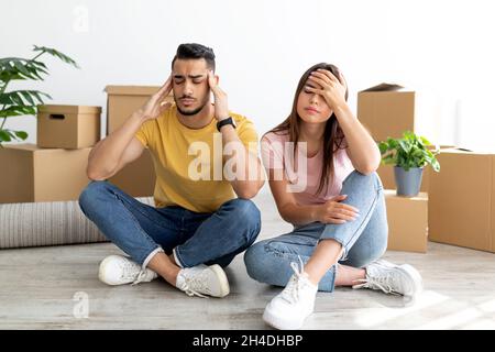 Tired young multiracial couple sitting on floor among boxes, massaging temples, exhausted from moving to new home Stock Photo